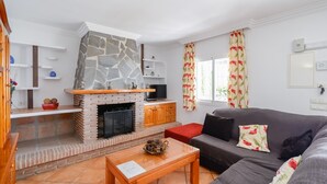 A living room with a stone fireplace, a grey sofa, wooden shelves, and a window with floral curtains. - Villa Pastora Nerja by Ruralidays (Málaga)