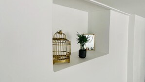 Un mur blanc sur lequel est fixée une étagère, et sur laquelle reposent une cage à oiseaux dorée et un pot de fleurs.