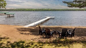 Uno scorcio di un lago con un molo che si protende sull'acqua, circondato da una spiaggia sabbiosa e diverse sedie Adirondack.
