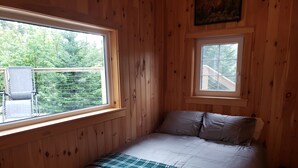 Een houten kamer met een bed, twee ramen en uitzicht op groen.