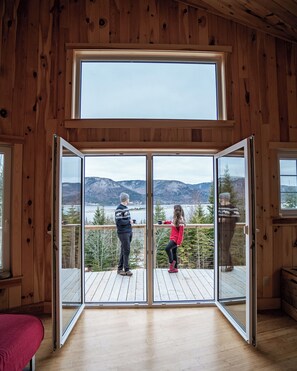 Una cabaña de madera con un amplio ventanal corredera que da a una terraza con vista a montañas y un lago.