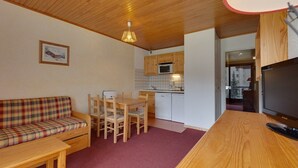 A cozy room with a wooden ceiling, a plaid sofa, a dining table with chairs, a kitchen area with white cabinets, and a television on a wooden stand.