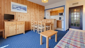 A cozy room with a wooden wall, a dining table with chairs, a bed, a TV, and a kitchen area with cabinets and a microwave.