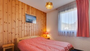 A bedroom with a wooden paneled wall, a bed with a red blanket, a nightstand with a lamp, a small wooden stool, and a window with curtains.