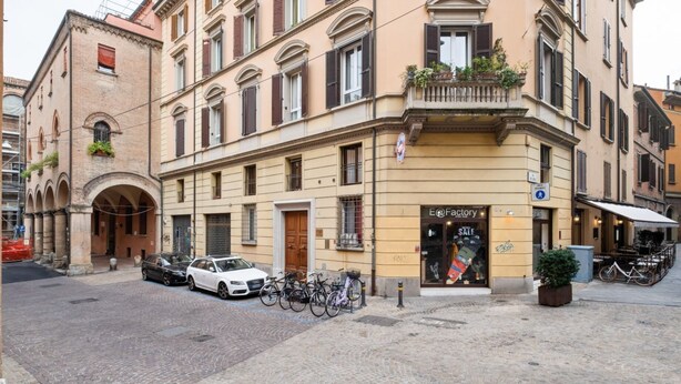 Un edificio con escaparate identificado como 'E@Factory' y un letrero de venta. Hay bicicletas estacionadas afuera, un coche blanco y una calle adoquinada.