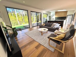 A modern living room with a sofa, coffee table, and chairs, large windows with a view of greenery, and a kitchen area.