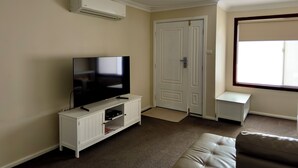 A living room with a flat-screen TV on a white cabinet, a beige couch, a white door, and a window with blinds.