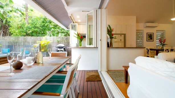 A modern kitchen with white cabinets, a dining area with a wooden table and chairs, and a view of lush greenery outside.