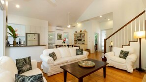 A spacious living area with a white sofa set, a coffee table, and a dining table. There is a kitchen area with white cabinets and a dining table with chairs. A staircase with a wooden railing is visible in the background.