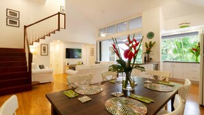 A modern living room with a wooden staircase, a dining area with tropical-themed tableware, and a kitchen with large windows.