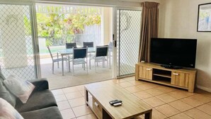 A living room with a sofa, a coffee table, a TV on a wooden stand, and a sliding glass door leading to an outdoor area with chairs and a table. - Standard Three-Bedroom Villa with Private Pool (Palm Cove)