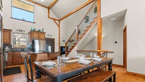A spacious dining area with a wooden table set for four, a staircase with wooden railings, and a kitchen in the background. - Pools Pvt Hot Tub near Thunder Ridge & Big Cedar (Hollister)