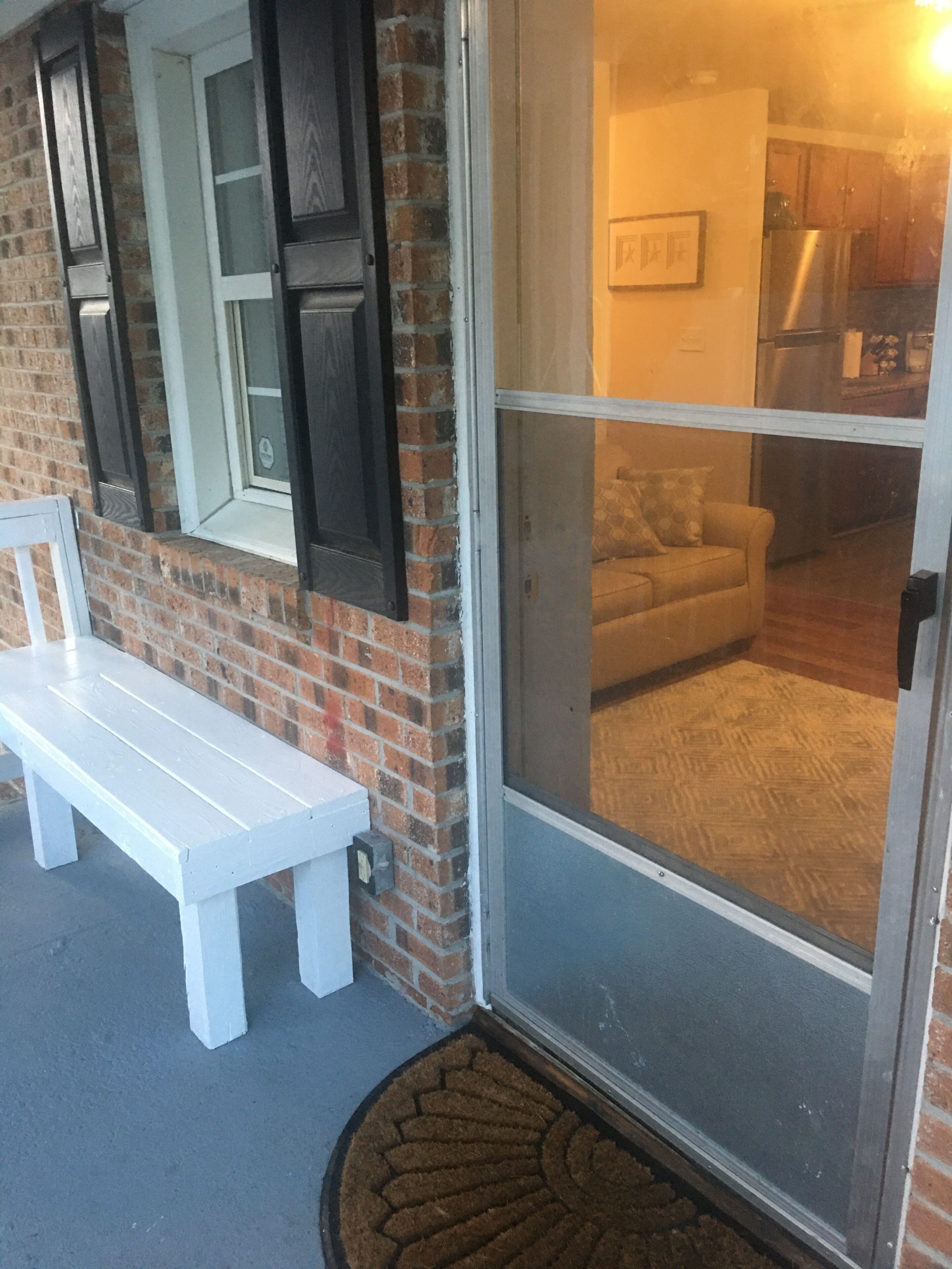A white bench outside a house with a brick wall and a sliding glass door leading to a living room with a sofa and a kitchen area visible through the door.