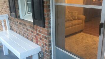A white bench outside a house with a brick wall and a sliding glass door leading to a living room with a sofa and a kitchen area visible through the door.