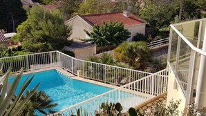 A pool with a white railing, surrounded by greenery and overlooking a coastal area with houses.