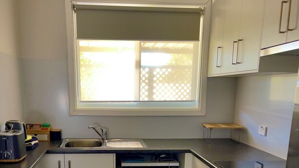 Una cocina moderna con armarios blancos, horno empotrado, microondas, pileta y ventana con persianas.