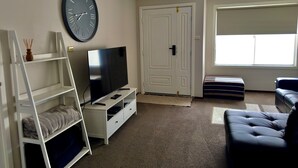 Sala de estar con televisor de pantalla plana, estante blanco, un reloj de pared grande, una puerta y una ventana.