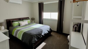 Un dormitorio con cama, ventilador de techo, ventana con cortinas, mesita de noche con lámpara y armario.