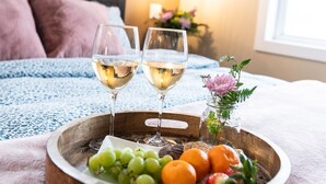 Una bandeja de madera con dos copas de vino blanco, uvas y naranjas sobre una cama con una manta azul.