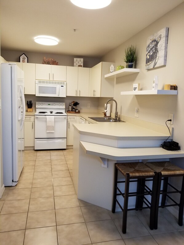 A modern kitchen with white cabinetry, a built-in oven, a microwave, and a sink. There are two stools at the island counter. - Cozy and spacious 3/2 condo. Family friendly. 3 Mins to the beach. Dog friendly (Naples)