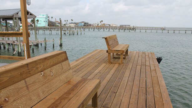 Un muelle de madera con bancos que se extiende sobre aguas tranquilas, una vista distante de un muelle con edificios, y un cielo nublado.