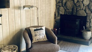 Un salon chaleureux avec une cheminée en pierre, un téléviseur fixé au mur et un fauteuil agrémenté d’un coussin aux motifs inspirants.