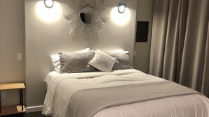 Un lit bien fait, avec une couette blanche et plusieurs oreillers, un luminaire fixé au mur et un sol carrelé foncé.