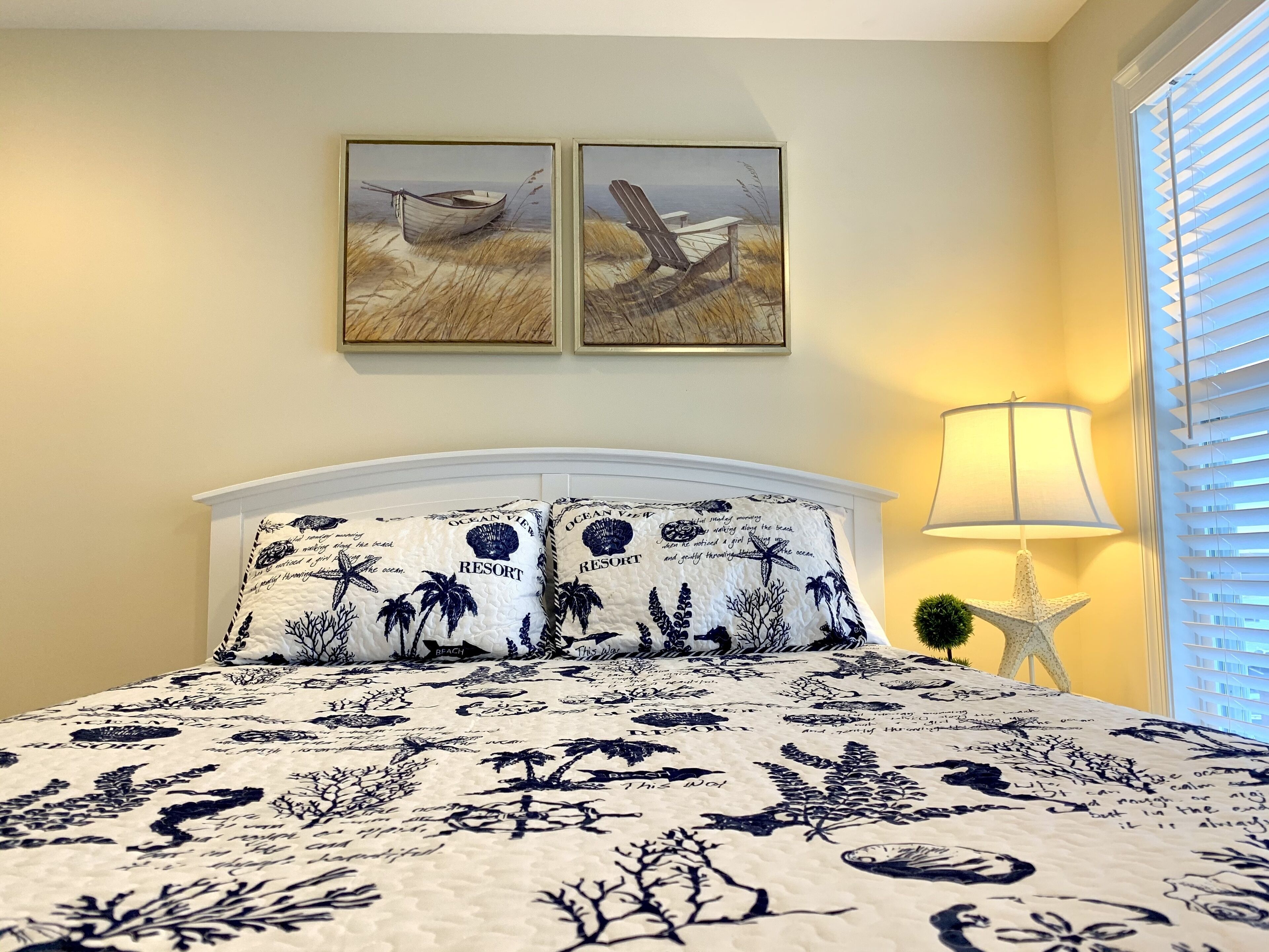 Quarto com cama, duas pinturas na parede, abajur e janela com persianas.