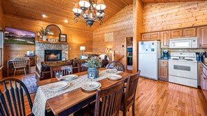 Interior acogedor de una cabaña de troncos, con comedor, cocina, chimenea y un cuadro colgado.