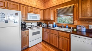 Una cocina con gabinetes de madera, un refrigerador blanco, un microondas, una estufa, un lavaplatos y una ventana con persianas.