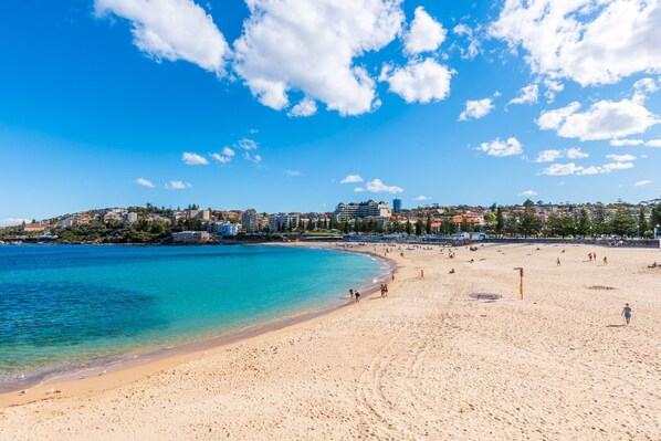 A beach with clear blue water, sandy shore, and buildings in the background.