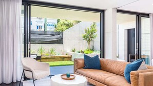 A modern living room with a leather sofa, a round coffee table, and a view of a garden and building in the background. - The Arden 1 Coogee New large Beachside Garden Apt (Sydney)
