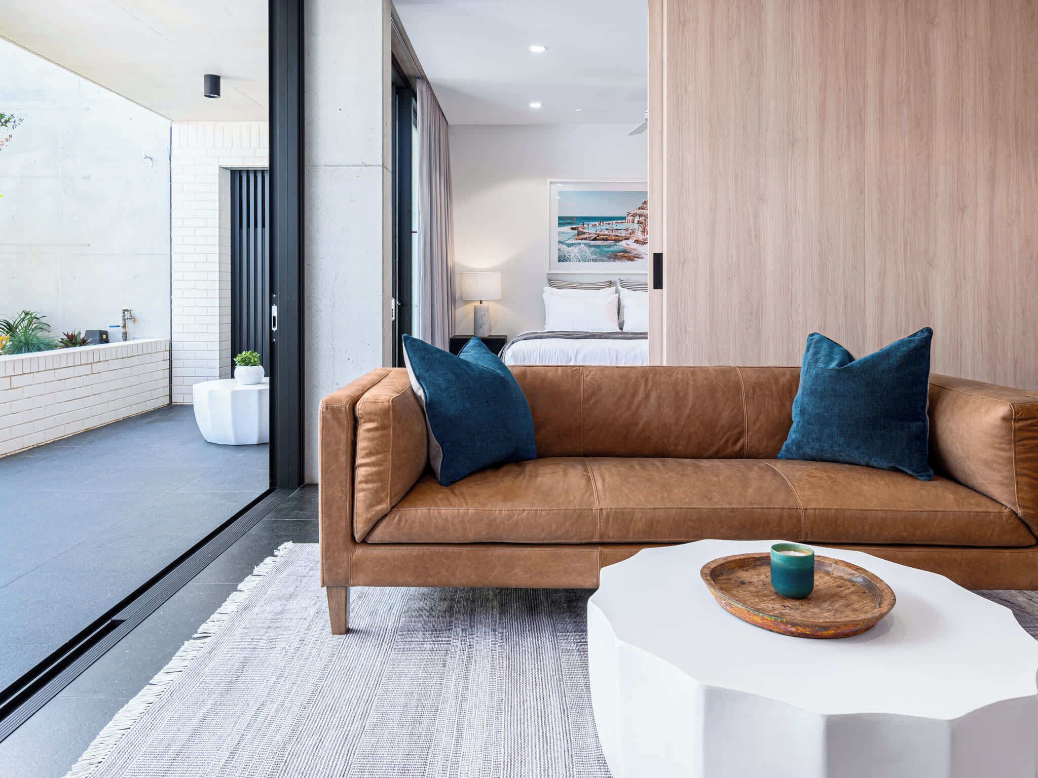 Uma sala de estar moderna com um sofá de couro marrom, duas almofadas azuis, uma mesa lateral branca com uma vela verde-água e vista para a praia através de uma janela grande.