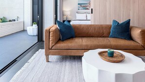 A modern living room with a brown leather sofa, two blue cushions, a white side table with a teal candle, and a view of the beach through a large window. - The Arden 1 Coogee New large Beachside Garden Apt (Sydney)