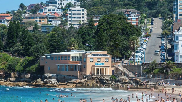 A beach with people sunbathing and swimming, a building with a clock, and a parking area with cars. - The Arden 1 Coogee New large Beachside Garden Apt (Sydney)