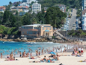 A beach with people sunbathing and swimming, a building with a clock, and a parking area with cars. - The Arden 1 Coogee New large Beachside Garden Apt (Sydney)