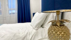 Una cama bien hecha con sábanas blancas, una lámpara dorada con forma de piña y un cabecero azul.