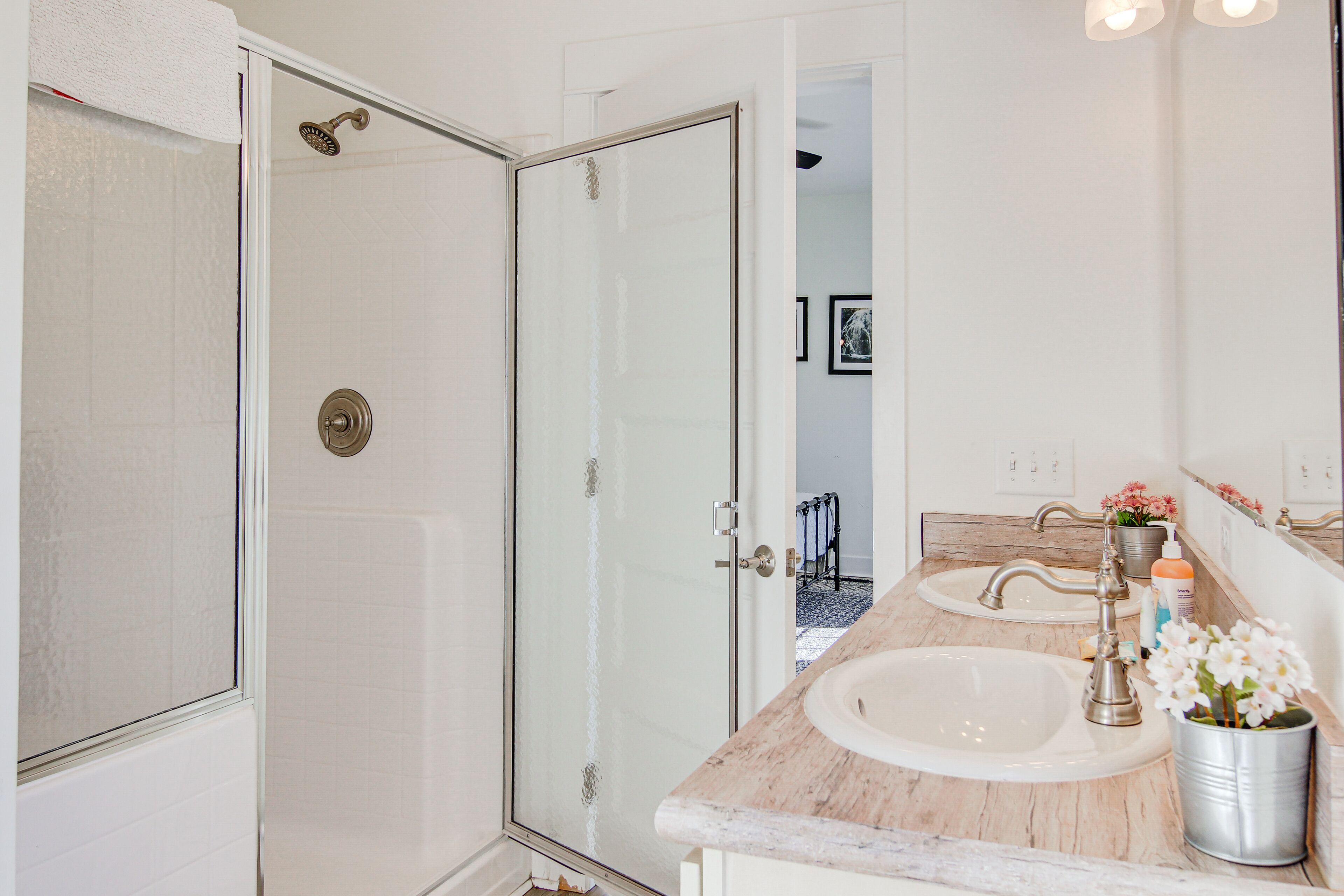 A bathroom with a glass shower door, a sink with a marble countertop, and a mirror with a flower vase.