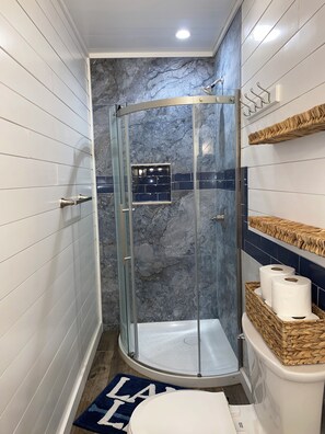 Un baño con ducha envuelta en vidrio, paredes de azulejos blancos, salpicadero de mármol y una alfombra azul.