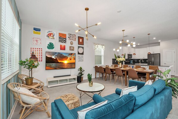 A modern living room with a blue sofa, dining table, and chairs, a kitchen area with appliances, and artwork on the walls. - Artsy 9BR Mansion for 24 people with 6 mil Disney w Pool&Water Park (Kissimmee)