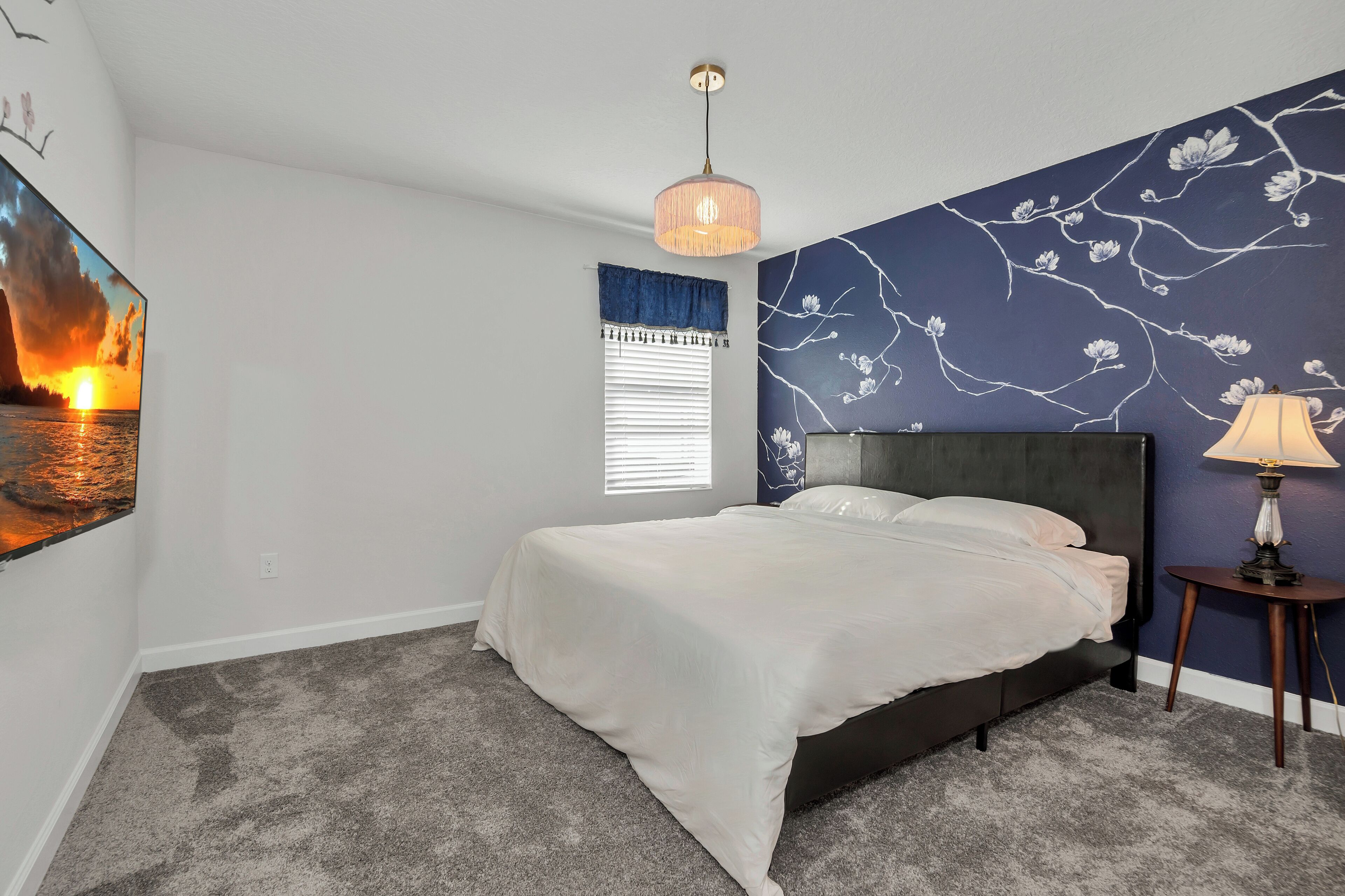 A bedroom with a bed, a nightstand, a lamp, a window with blinds, and a wall with a floral mural.