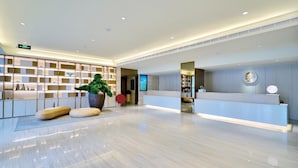 A modern hotel lobby with a reception desk, a large potted plant, and a wall-mounted digital screen.