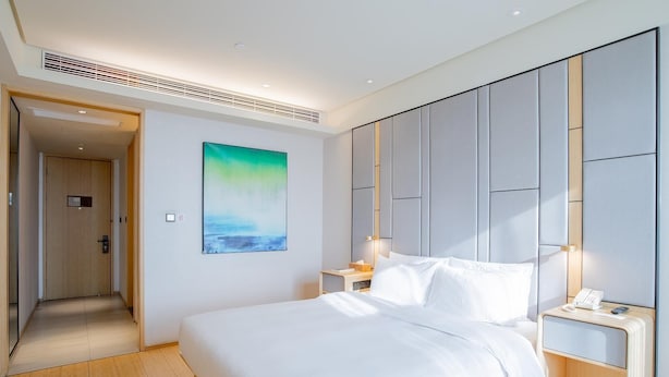 A modern hotel room with a large bed, a nightstand, a wall-mounted artwork, and a door leading to another room.