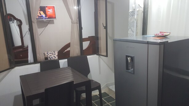 Una habitación compacta con una mesita y sillas, un refrigerador, un televisor montado en la pared y una ventana con cortinas.