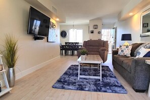 A living room with a sofa, a coffee table, a TV mounted on the wall, and a dining area with chairs and a table.