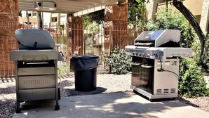 Una griglia per barbecue e un distributore d'acqua in un'area esterna con pavimento in cemento, circondata da una recinzione e vegetazione.