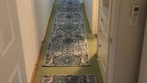 Ein schmaler Flur mit einem gemusterten Teppich, einem Stuhl im Hintergrund und einer Tür rechts.