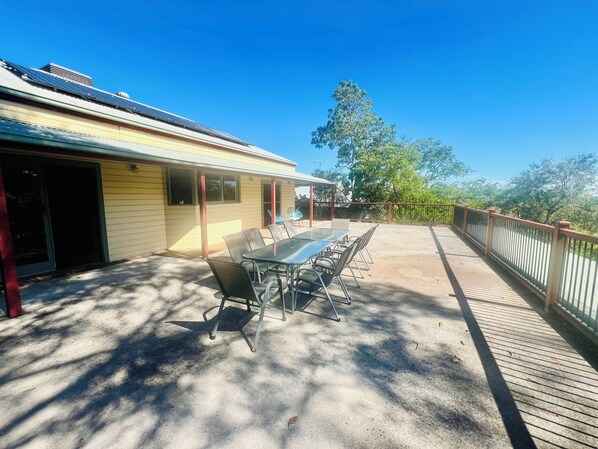 A covered patio with a table and chairs, a yellow building with solar panels, and a view of trees.