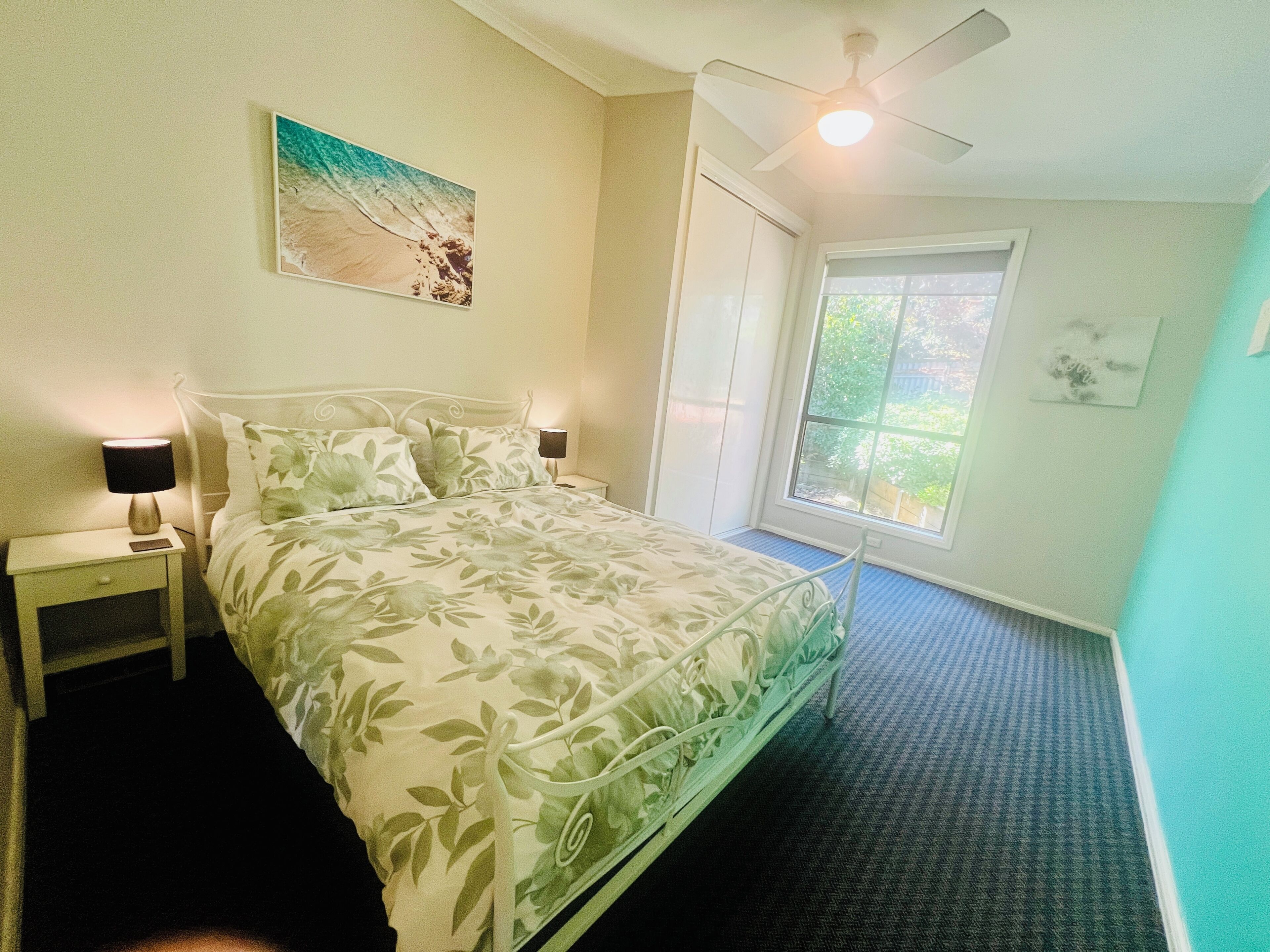 Une chambre à coucher comprenant un lit, des tables de chevet, un ventilateur de plafond, une fenêtre avec vue et un tableau accroché au mur.