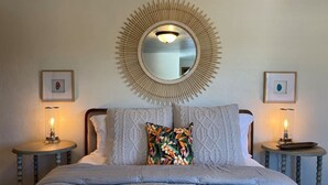 Ein Schlafzimmer mit einem großen Bett, einem runden Spiegel mit Sonnenspeiche-Rahmen, zwei Nachttischen mit Lampen und gerahmten Bildern an den Wänden.